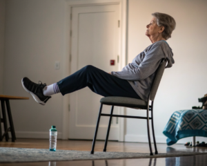 Persona mayor genérica sentada en una silla sin brazos realizando una elevación de talones, manos en los muslos, entorno de rehabilitación en casa con cojín y botella; enfoque cerrado en torso, manos y pies, luz suave y fondo desenfocado.