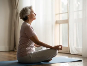 Mujer mayor realizando estiramientos de yoga en una habitación iluminada suavemente, simbolizando la paz y el bienestar en la tercera edad.