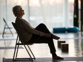 Senior practicando yoga adaptado en una silla en un entorno tranquilo con iluminación suave. La imagen destaca la postura del senior y la textura de la silla, con un fondo desenfocado que incluye elementos de yoga.