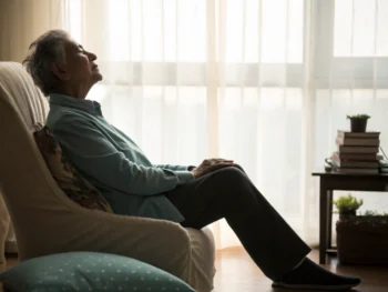 Adulto mayor practicando progresiva relajación muscular en un ambiente sereno y suave, sentado en una silla cómoda con sus manos en el regazo.