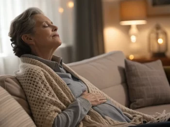 Anciano practicando respiración consciente en un salón acogedor, con la mano en el abdomen y ojos cerrados, rodeado de luz suave y texturas de manta.